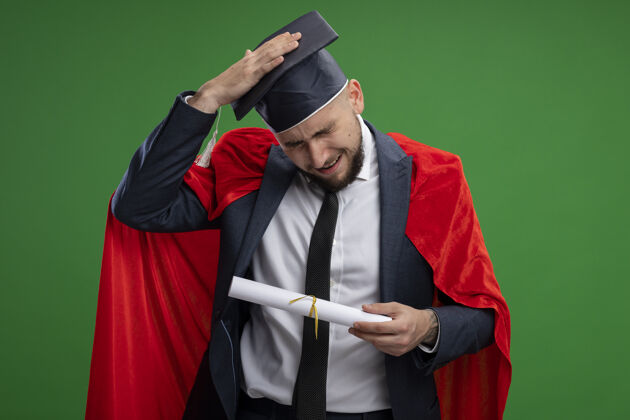 错误一个身穿红斗篷的毕业男子拿着毕业证书 头上戴着一只手 看起来很困惑 因为他站在绿色的墙上犯了错误抱毕业困惑