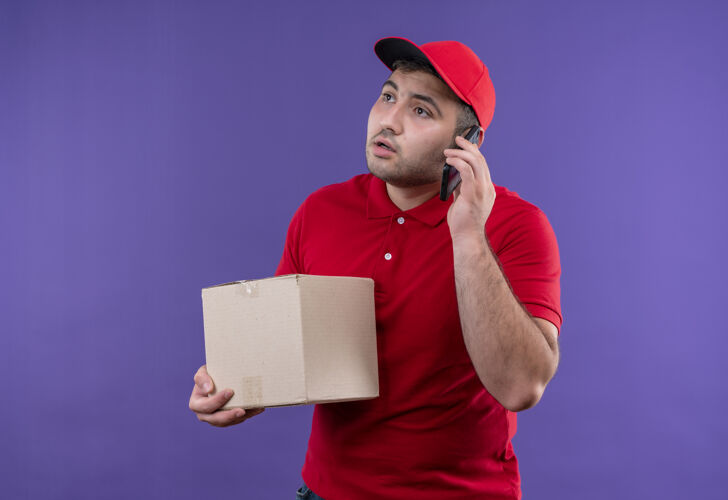 年轻站在紫色的墙上 一个身穿红色制服 戴着帽子 拿着盒子的年轻送货员严肃地一边看一边讲着手机送货人人