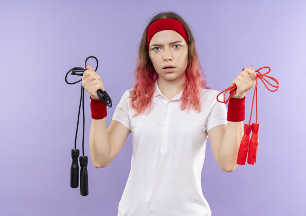 绳子戴着头巾 拿着跳绳 困惑地站在紫色墙壁上的年轻运动女郎市民姿势困惑