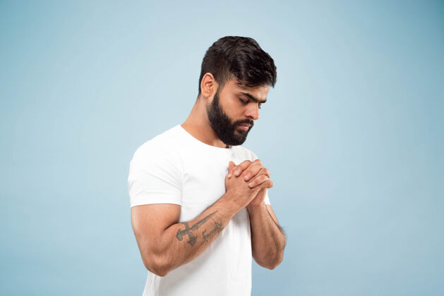 英俊半身特写照片 是一个穿着白衬衫的印地安人 隔离在蓝色背景上人类的情感 面部表情 广告概念负面空间闭着眼睛站着祈祷祈祷封闭表情