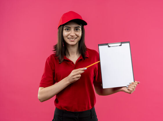 铅笔穿着红色马球衫 头戴帽子的年轻送货女孩拿着一本空白的笔记本 用铅笔指着笔记本 微笑着站在粉色背景上 脸上洋溢着幸福衬衫页面指向