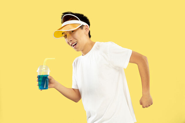 帽子黄色工作室背景上的韩国年轻人半身像穿着白衬衫 戴着黄色帽子的男模喝鸡尾酒人类情感的概念 表达 夏天 假期 周末年轻人惊奇