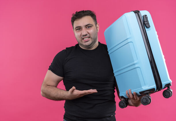 旅行身穿黑色t恤的年轻旅行家拿着手提箱站在粉红色的墙上 用手挽着手提箱 脸上带着恼怒的表情假期姿势旅行者