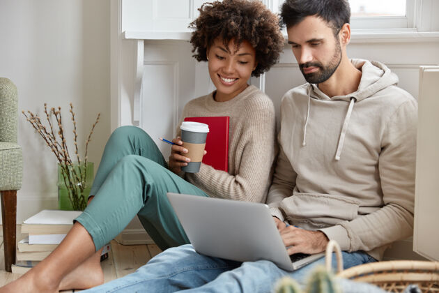快乐浪漫的情侣们坐在地板上 专注于笔记本电脑 在线观看有趣的电影 享受芳香的咖啡 心情愉快 享受无线网络连接 有空余时间休闲室内女人