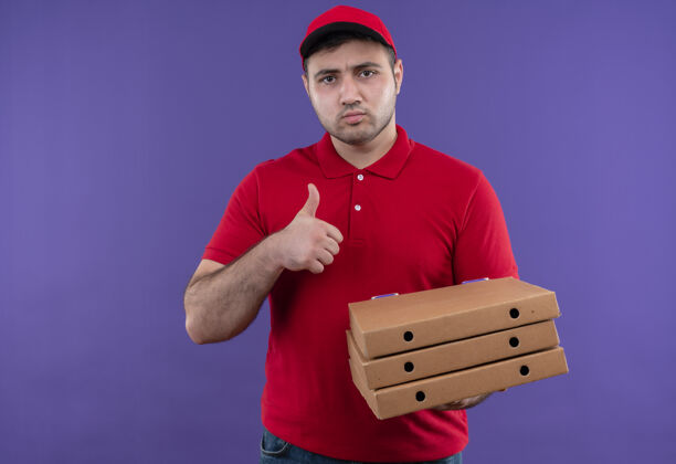 送货年轻的送货员身穿红色制服 戴着帽子 手里拿着披萨盒 皱着眉头 竖起大拇指站在紫色的墙上站着帽子市民