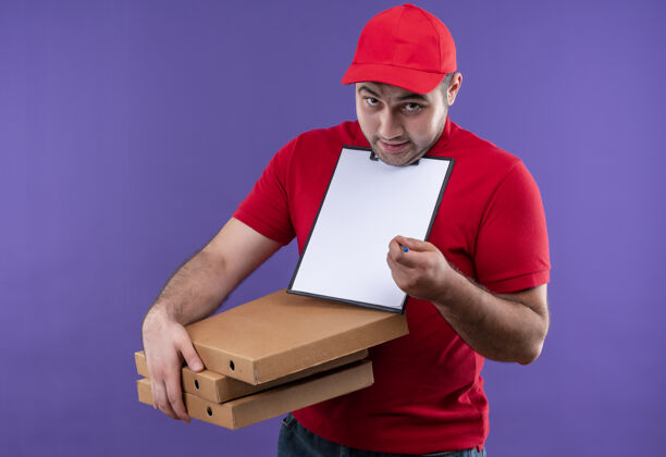 签名身穿红色制服 头戴鸭舌帽的年轻送货员站在紫色的墙上 手里拿着披萨盒和空白页的剪贴板 困惑地微笑着要求签名页面年轻人空白