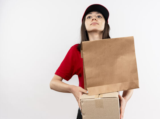 帽子身穿红色制服 头戴鸭舌帽 手拿纸包和纸箱的年轻送货员站在白色背景下 自信地看着镜头女孩交货举行