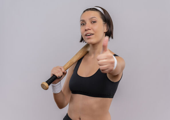 表演身穿运动服的年轻健身女士手持棒球棒 自信地微笑着竖起大拇指站在白色的墙上运动员人自信