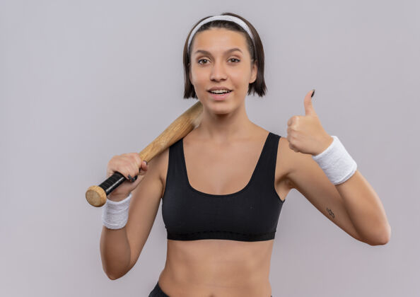 棒球身穿运动服的年轻健身女士手持棒球棒 自信地微笑着竖起大拇指站在白色的墙上姿势运动运动员