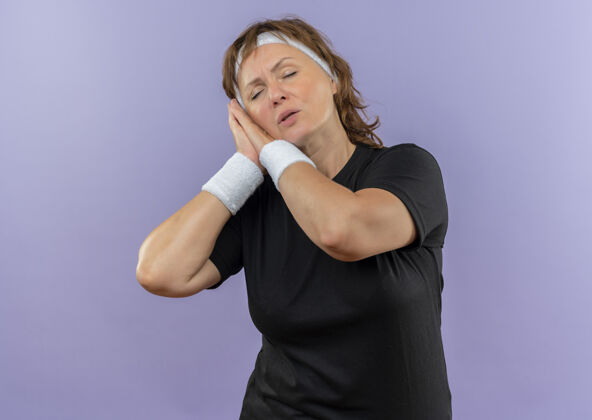 中间中年运动女性 身穿黑色t恤 头带 双手合十 头靠在手心上 眼睛闭着 想要站在蓝色的墙上睡觉年龄眼睛睡眠
