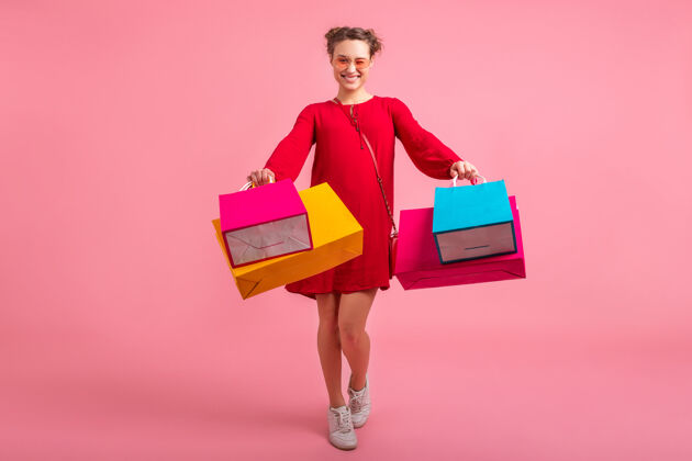 夏天迷人的快乐微笑的时尚女性购物狂穿着红色的新潮连衣裙拿着五颜六色的购物袋在粉色的墙上孤立着 销售兴奋 时尚潮流颜色购物狂休闲