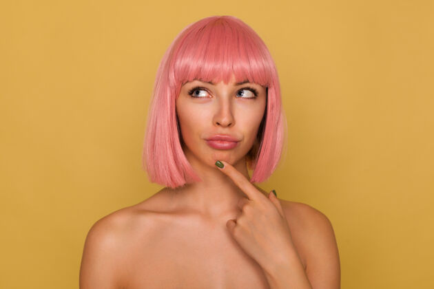 肖像年轻迷人的粉红色头发的女性 鲍勃式的发型沉思地向上看 双唇合拢 食指放在下巴上 站在芥末墙上食指邦化妆