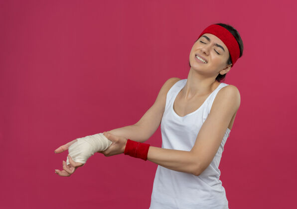 女人穿着运动服的年轻健身女 戴着头巾 看起来不舒服 摸着缠着绷带的手腕 站在粉红色的墙上感到疼痛长相姿势触摸