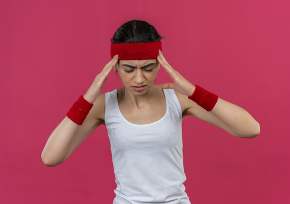 运动穿着运动服的年轻健身女士戴着头巾 站在粉色的墙上摸着她的太阳穴 头痛得要命健身头痛健康