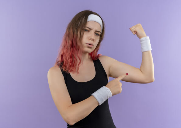 展示身穿运动服的年轻健身女士举起拳头 站在紫色的墙上 露出自信的二头肌健身姿势运动