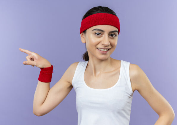 微笑身穿运动服的年轻健身女士戴着头巾 微笑着 用食指指着站在紫色墙上的一边运动目录指点