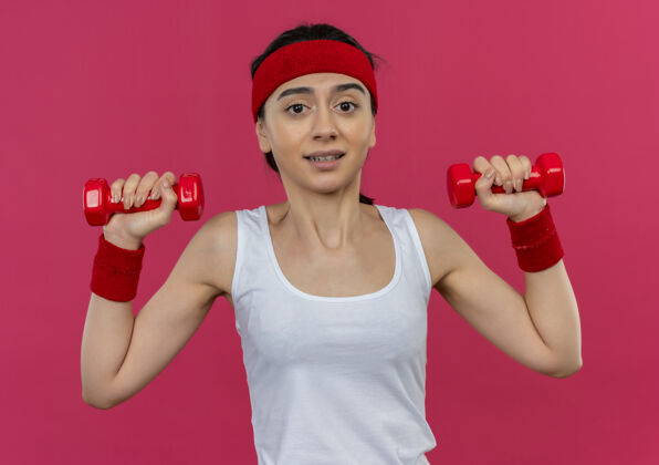 女人身穿运动服 头箍 双手举着两个哑铃的年轻健身女士站在粉色的墙上市民手看