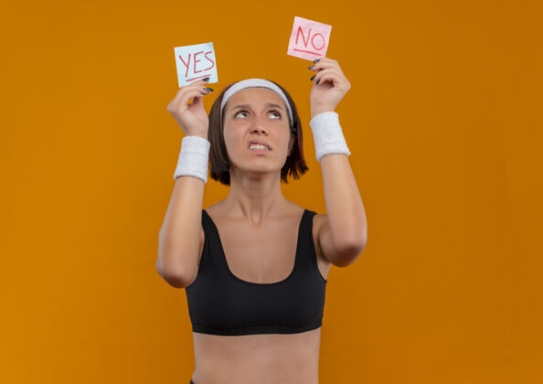 姿势穿着运动服的年轻健身女 头上戴着头巾 举着两张写着“是”和“否”的提醒纸 站在橘色的墙上困惑地看着它们单词运动人