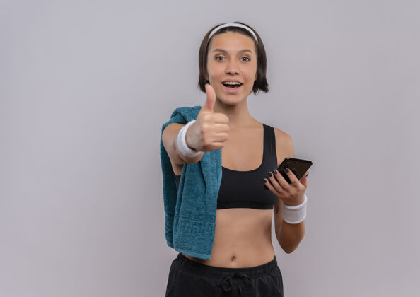 站着身穿运动服的年轻健身女士 肩上挎着毛巾 手持智能手机 微笑着竖起大拇指站在白色的墙上女人移动女人