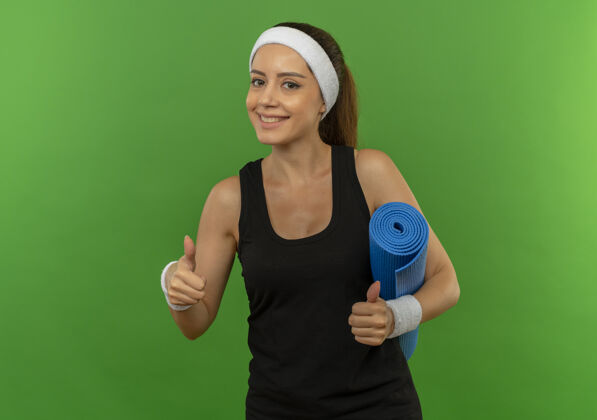 瑜伽身穿运动服 头戴瑜伽垫 竖起大拇指 微笑自信地站在绿色墙壁上的年轻健身女士健身运动表演