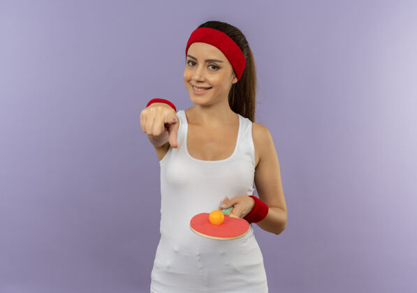 网球身穿运动服的年轻健身女 头戴头巾 手持球拍和乒乓球 用食指指着镜头 脸上带着微笑站在灰色的墙上站着市动