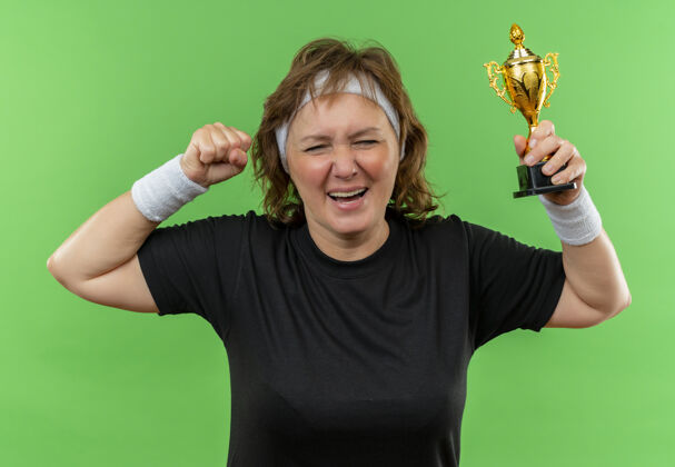 快乐中年运动女性 身穿黑色t恤 头戴头巾 手持奖杯 握紧拳头 站在绿色的墙上 快乐而兴奋运动装年龄拳头