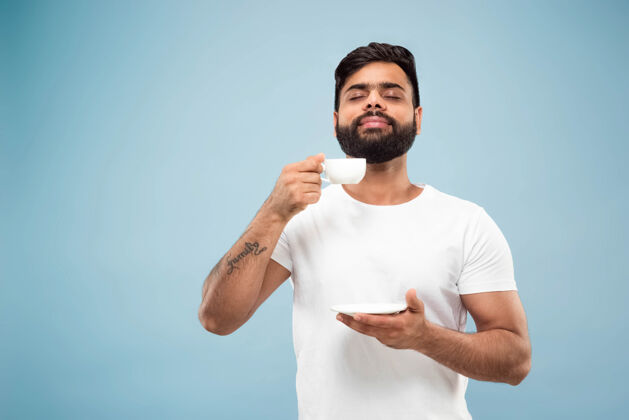 就业蓝色背景上穿着白衬衫的印度年轻人的半身特写肖像人类情感 面部表情 销售 广告概念负面空间喝咖啡或茶的享受男人成功长相