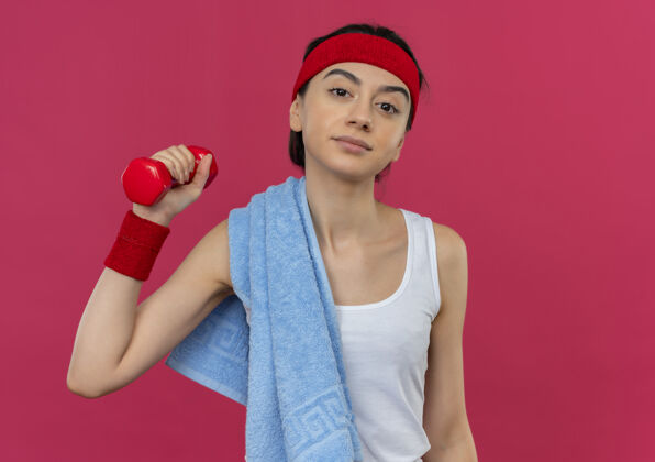 站身穿运动服的年轻健身女士 肩上戴着头带和毛巾 举起手拿着哑铃 站在粉色的墙上显得很自信看提高信心