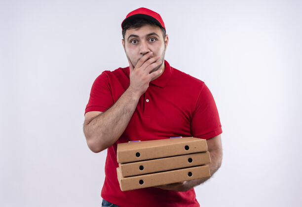 制服年轻的送货员身穿红色制服 戴着帽子 手里拿着披萨盒 微笑着 用手捂住嘴巴站在白色的墙上嘴拿着人