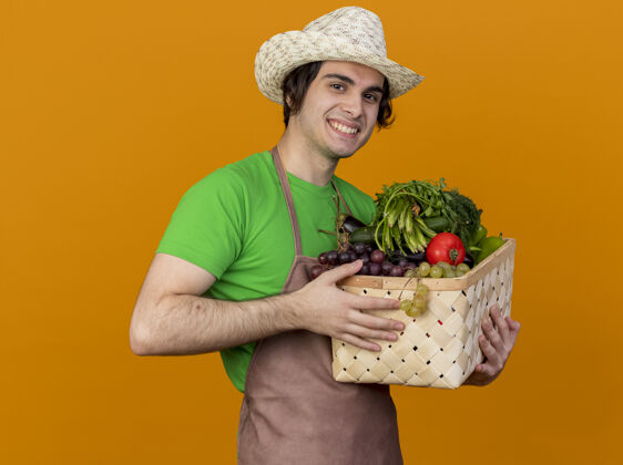 花园年轻的园丁 围着围裙 戴着帽子 手里拿着装满蔬菜的箱子 站在橙色的背景上 面带微笑地看着摄像机年轻立场蔬菜