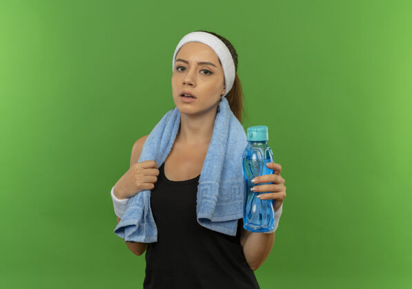 人一位身穿运动服的年轻健身女士 头上戴着头巾 脖子上挂着毛巾 手里拿着一瓶水 站在绿色的墙上 看上去很疲惫头带水累了