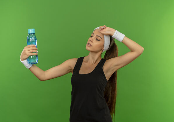 健康穿着运动服的年轻健身女 戴着头巾 手里拿着一瓶水 站在绿色的墙上 看上去很累女人瓶子运动