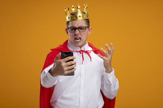 站着穿着红色斗篷戴着皇冠的超级英雄商人举着智能手机看着它 愤怒和沮丧地站在橙色背景上超级英雄商人皇冠