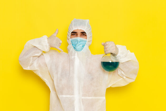 科学正面图穿着特殊防护服的男科学工作者拿着浅黄色表面上有蓝色溶液的烧瓶医学工人男性