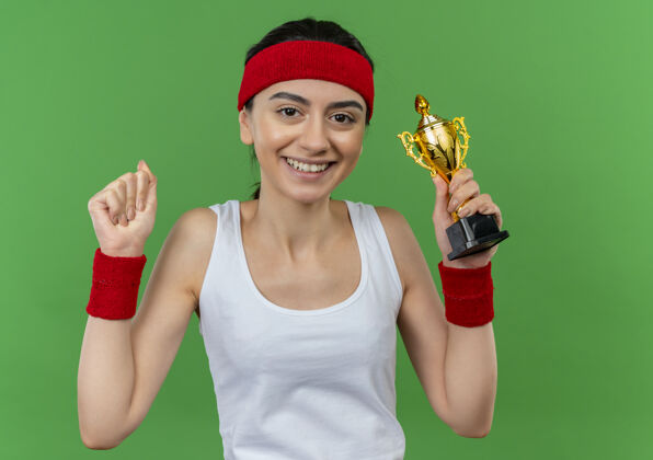 运动员身穿运动服的年轻健身女士 头箍高举拳头 像一个胜利者 微笑着自信地站在绿色的墙上喜欢提高微笑