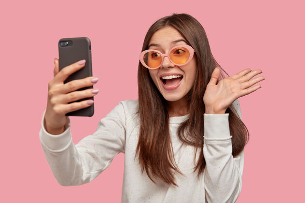 采取快乐的黑发女人戴着墨镜 打视频电话 连接无线网络 穿着白色套头衫 隔着粉红色的墙你好 朋友设备她自己模型