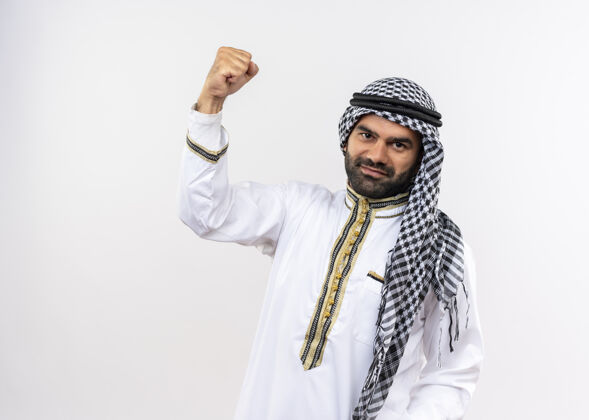 站身着传统服饰的阿拉伯男子高举拳头 微笑自信地站在白墙上阿拉伯语提高传统