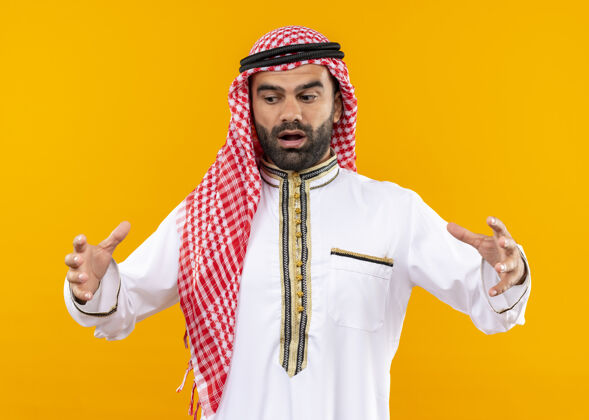 惊喜身着传统服装的阿拉伯商人用手展示大尺寸手势 测量符号 站在橙色的墙上显得很惊讶显示测量手