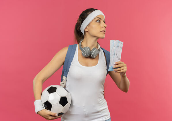 票一位身穿运动服 头戴头带 头戴耳机 手持机票和足球的年轻健身女士站在粉红色的墙上 严肃地看着一旁旁边年轻人持有