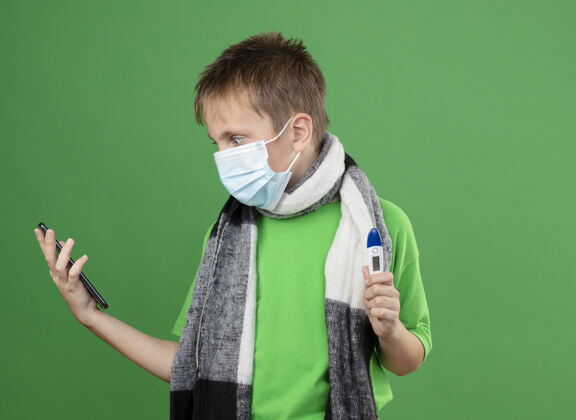 男孩生病的小男孩穿着绿色t恤 脖子上围着暖和的围巾 戴着护面面具 手里拿着温度计 站在绿色背景下焦急地看着他的智能手机站着温暖面部