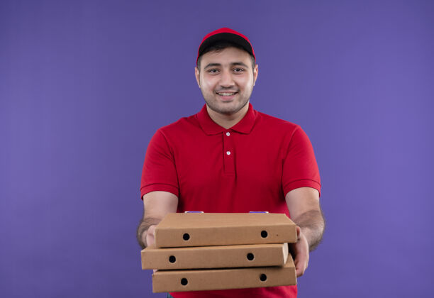 欢呼年轻的送货员身穿红色制服 戴着帽子 手里拿着披萨盒 站在紫色的墙上 开心地微笑着市民看着拿着