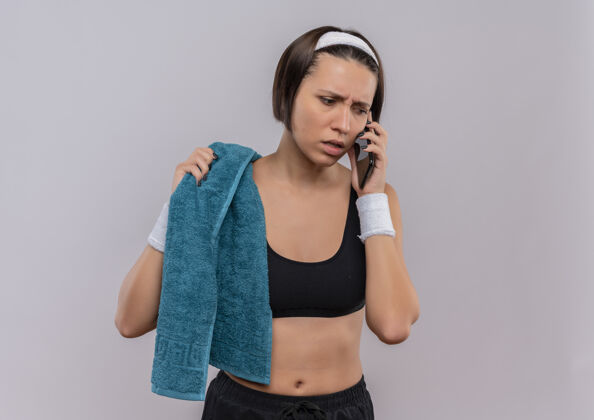 市民身穿运动服 肩上扛着毛巾的年轻健身女士站在白墙边讲手机 神情迷茫 焦虑运动女性困惑