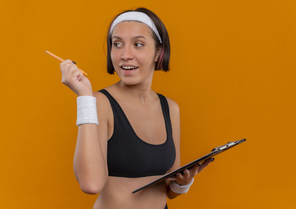 女人身穿运动服的年轻健身女 头上戴着头巾 手里拿着空白页的剪贴板 手里拿着一支笔 微笑着站在橙色的墙上微笑女性运动装
