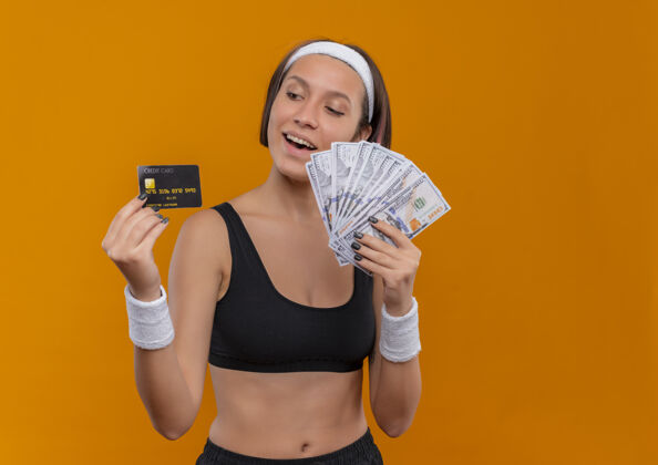 运动身穿运动服 头箍 手持信用卡 面带微笑的年轻健身女士站在橙色的墙上欢呼表演运动员