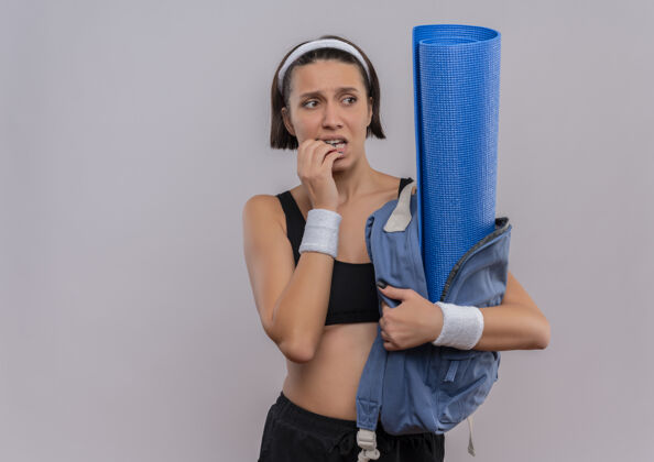 看穿着运动服的年轻健身女士背着背包 拿着瑜伽垫 看着一旁 紧张紧张地咬着白墙上的钉子站年轻人人