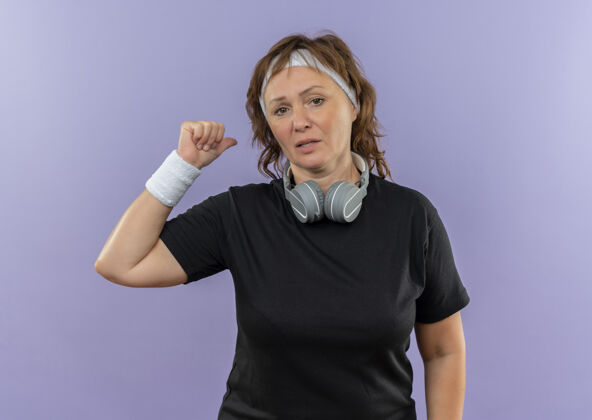 市民中年运动女性 身穿黑色t恤 头戴头带 手指向后 站在蓝色的墙上运动装中间头带