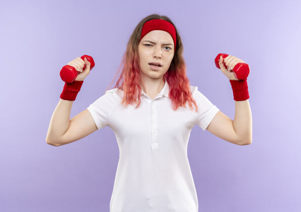 哑铃年轻的健身女士站在紫色的墙上用哑铃练习人脸姿势