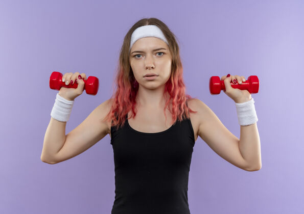 哑铃穿着运动服的年轻健身女士站在紫色的墙上 严肃地用哑铃做练习健身女人站立