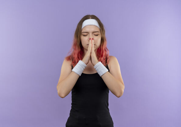运动身穿运动服的年轻健身女士双手合十 闭着眼睛祈祷 带着希望的表情站在紫色的墙上女人人立场