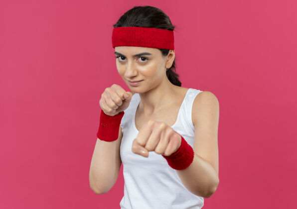 拳击手身穿运动服 头戴花边 微笑着的年轻健身女士 站在粉红色的墙上 摆出拳击手的姿势健身喜欢运动装